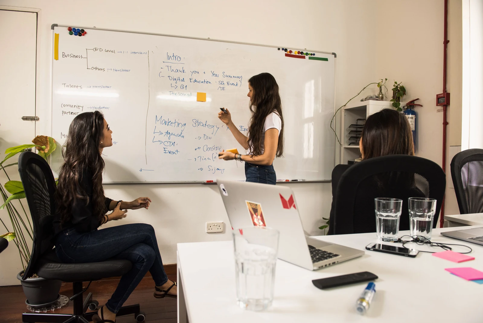 One colleague writes a plan on an office whiteboard while two teammates follow along from the table