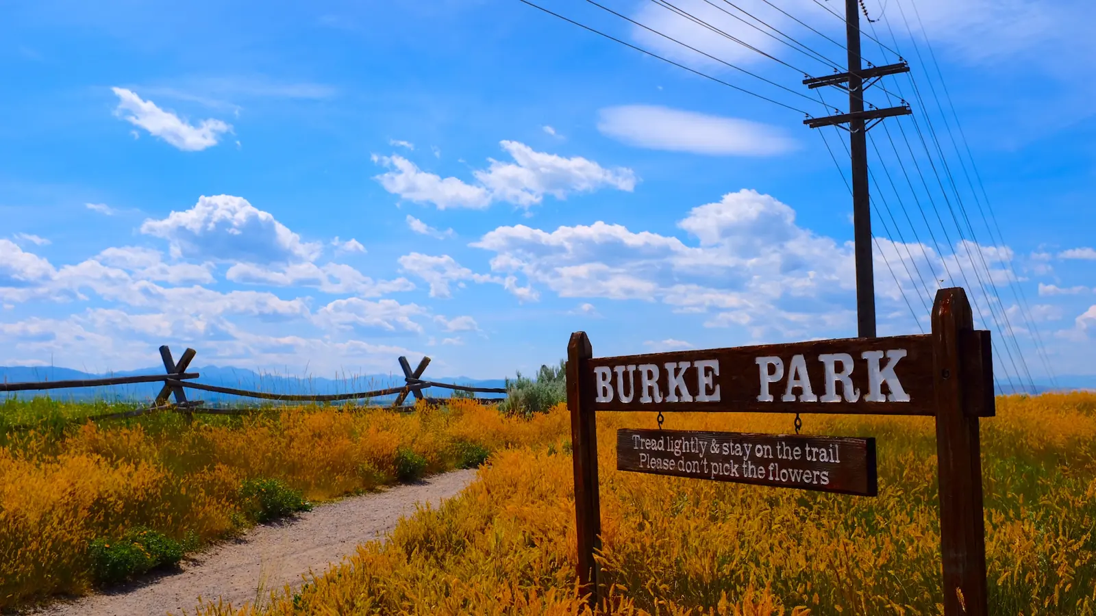 Burke Park trail in Bozeman, Montana A wooden Burke Park sign beside a gravel trail through yellow wildflowers, with a rail fence, a power pole and distant mountains under a blue sky