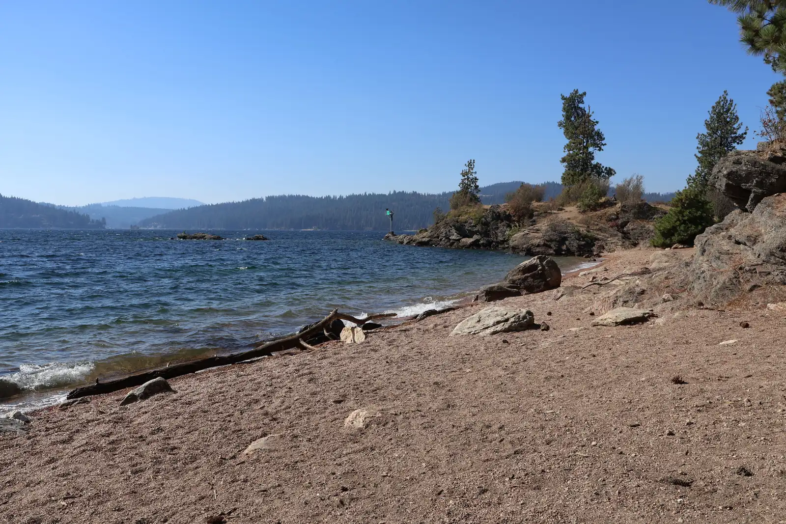 Lake Coeur d'Alene shoreline A sandy, rocky beach on Lake Coeur d'Alene with a driftwood log, a rocky point with pine trees and forested hills across the water