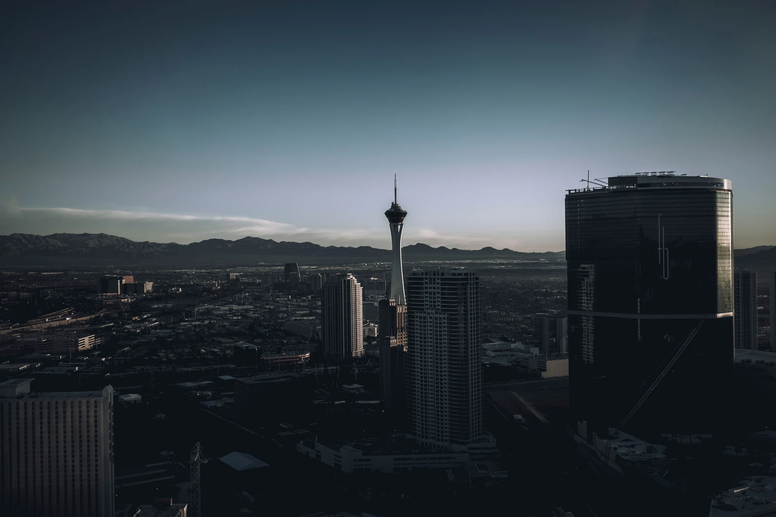 Elevated view over Las Vegas at dusk, with the STRAT tower and high-rise hotels in front of the city grid and a mountain range on the horizon