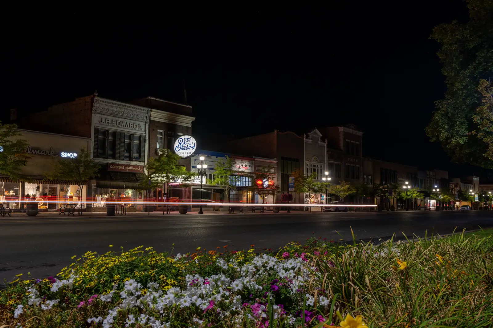 Historic brick storefronts on Main Street in downtown Logan at night, including the J.R. Edwards building and the lit Bluebird sign, with a flower bed in the foreground