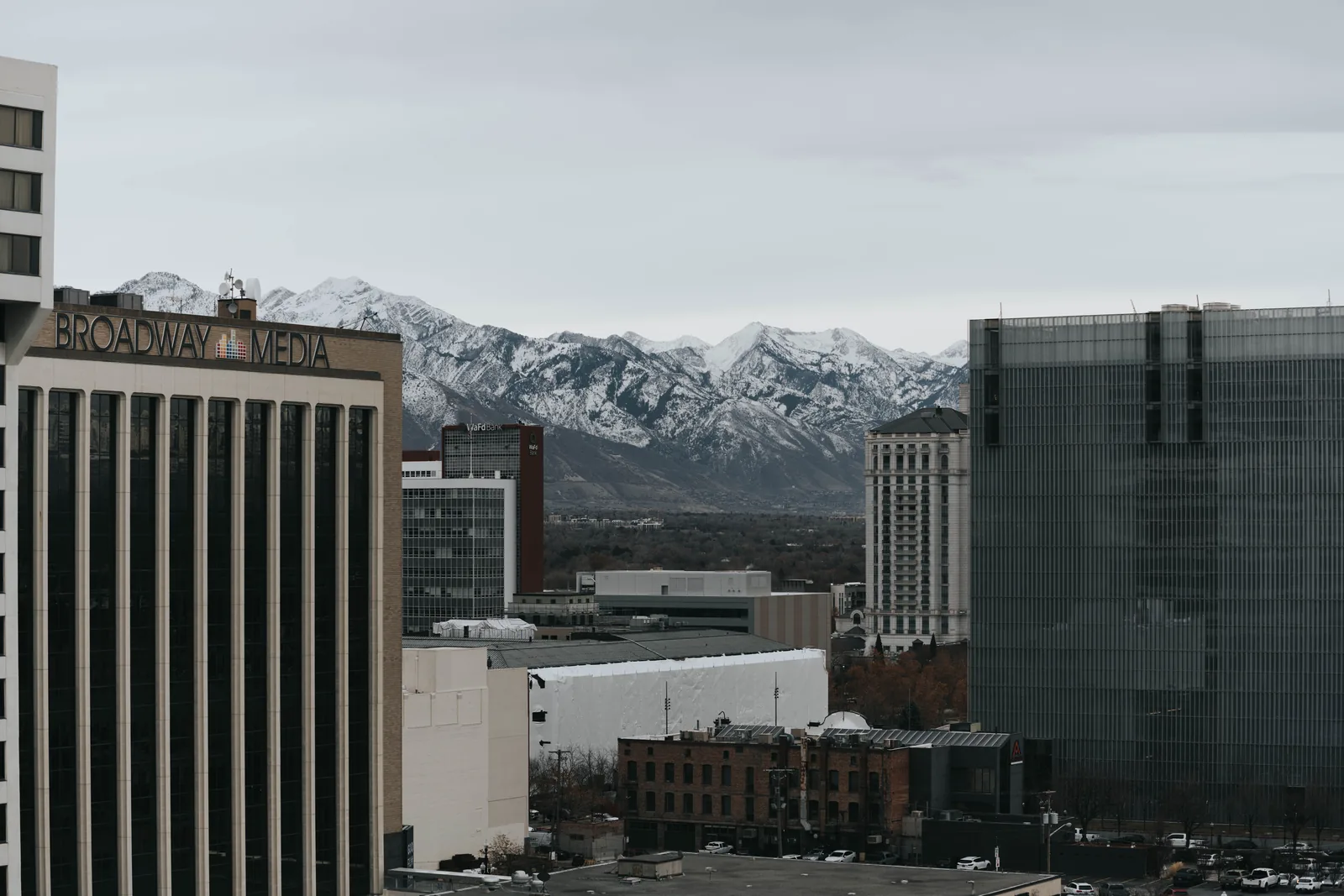 Downtown Salt Lake City office and apartment towers, including the Broadway Media building, with the snow-covered Wasatch Mountains behind under an overcast sky