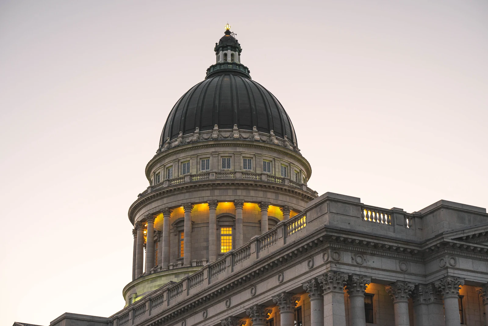 The lit dome and stone columns of the Utah State Capitol against a pale evening sky