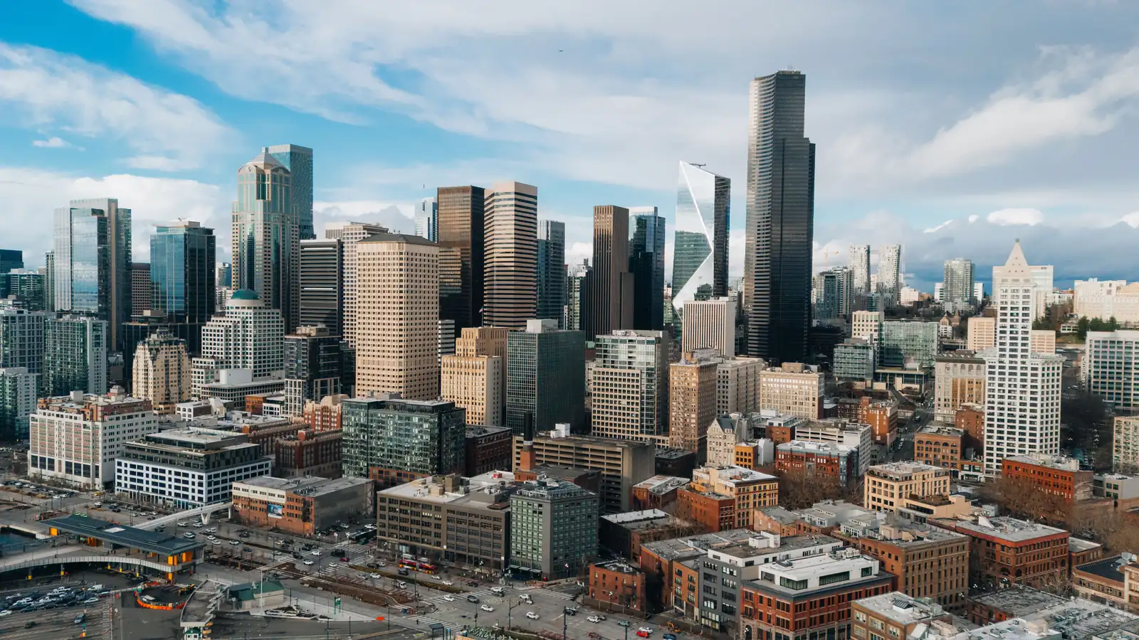 Aerial view of downtown Seattle's high-rise towers and older brick buildings, with rail tracks and a parking area in the foreground