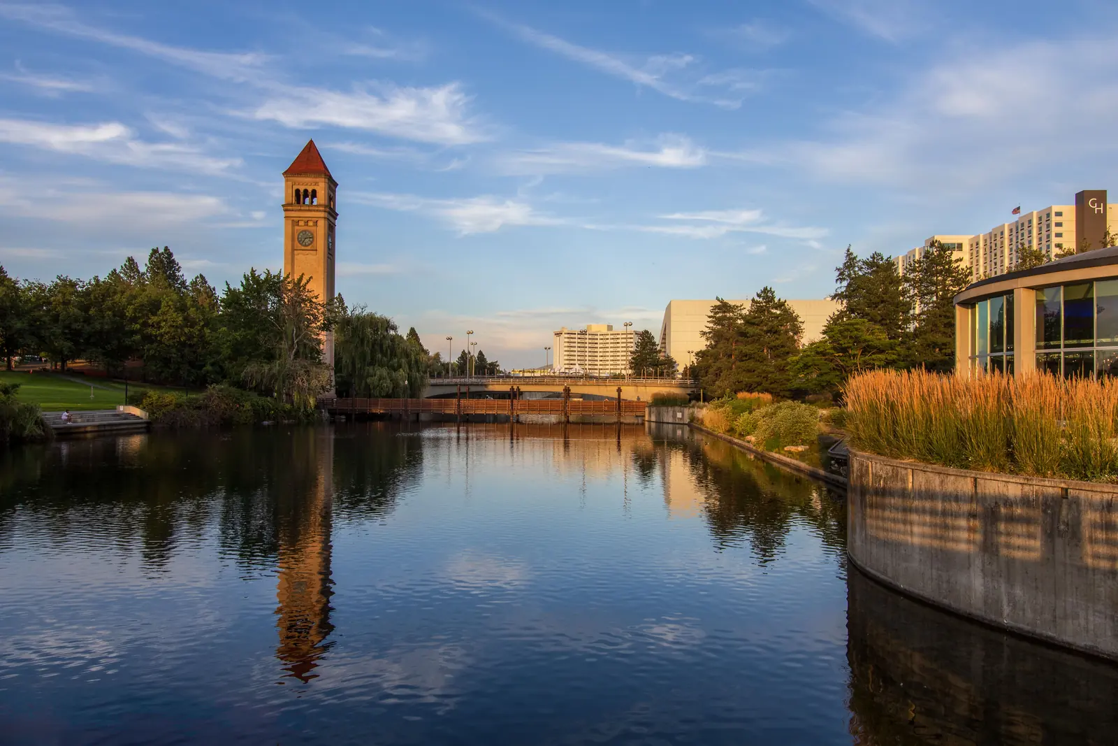The brick clock tower on the Spokane riverfront reflected in still water, with a footbridge, trees and downtown hotel buildings behind