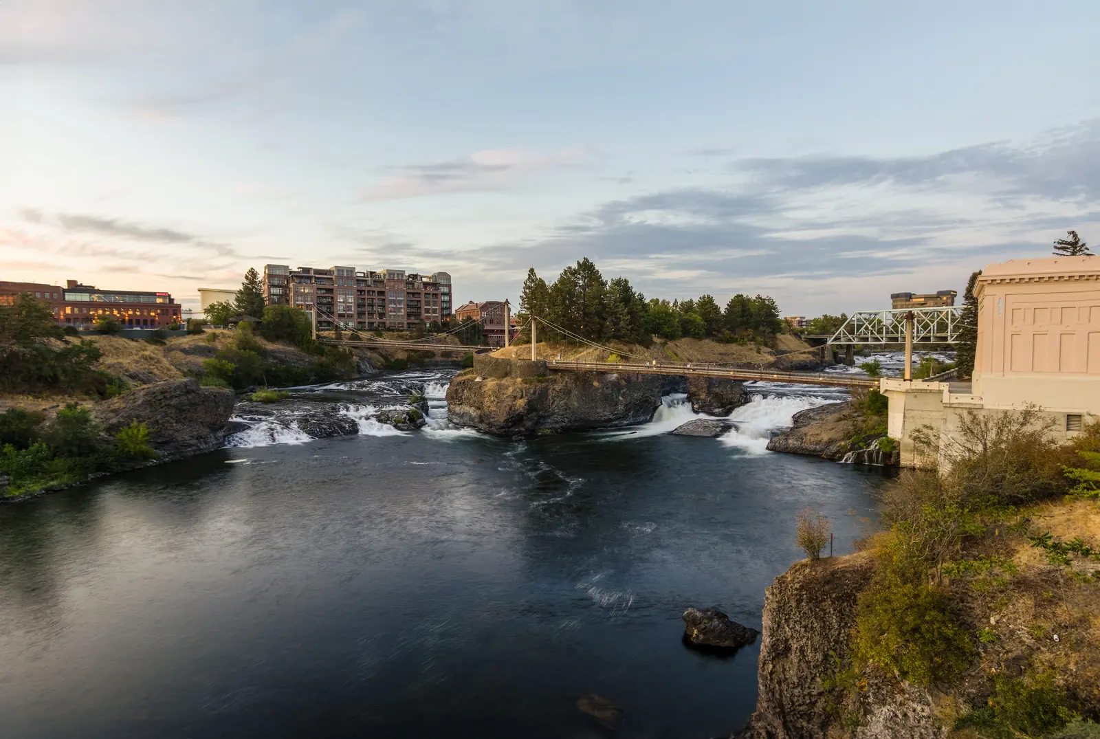 The Spokane River pooling below Spokane Falls, with a footbridge, a steel truss bridge and downtown Spokane apartment and office buildings at sunset