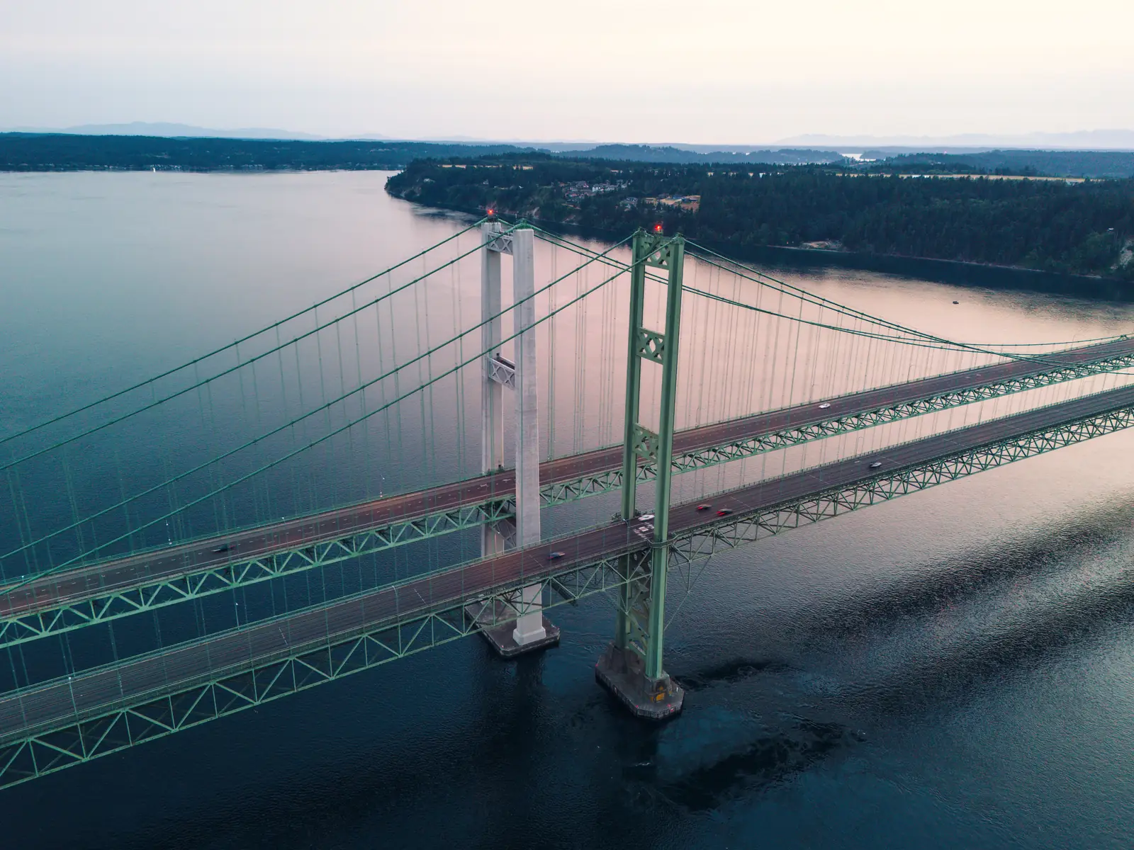 Aerial view of the twin green suspension spans of the Tacoma Narrows Bridge over calm water at dusk, with a forested shoreline behind