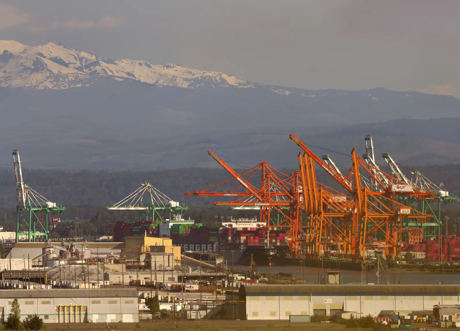 Orange and green container cranes and a loaded cargo ship at the Port of Tacoma, with warehouses in front and snow-capped mountains behind