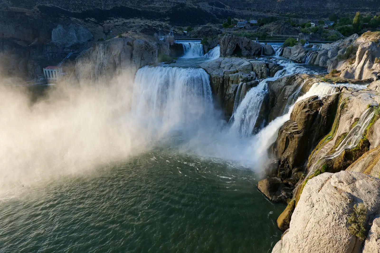 Shoshone Falls pouring over grey basalt cliffs into the green Snake River, with mist rising on the left and homes on the canyon rim behind